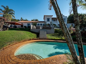 Ein Swimmingpool mit Ziegelrand, klares blaues Wasser und ein Haus im Hintergrund mit rotem Dach.