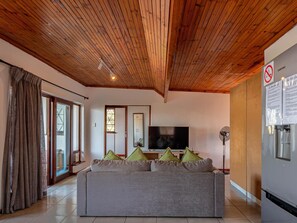 Ein Wohnzimmer mit Holzboden, einem grauen Sofa mit Kissen, einem Flachbildfernseher und einem Samsung-Kühlschrank an der Wand.