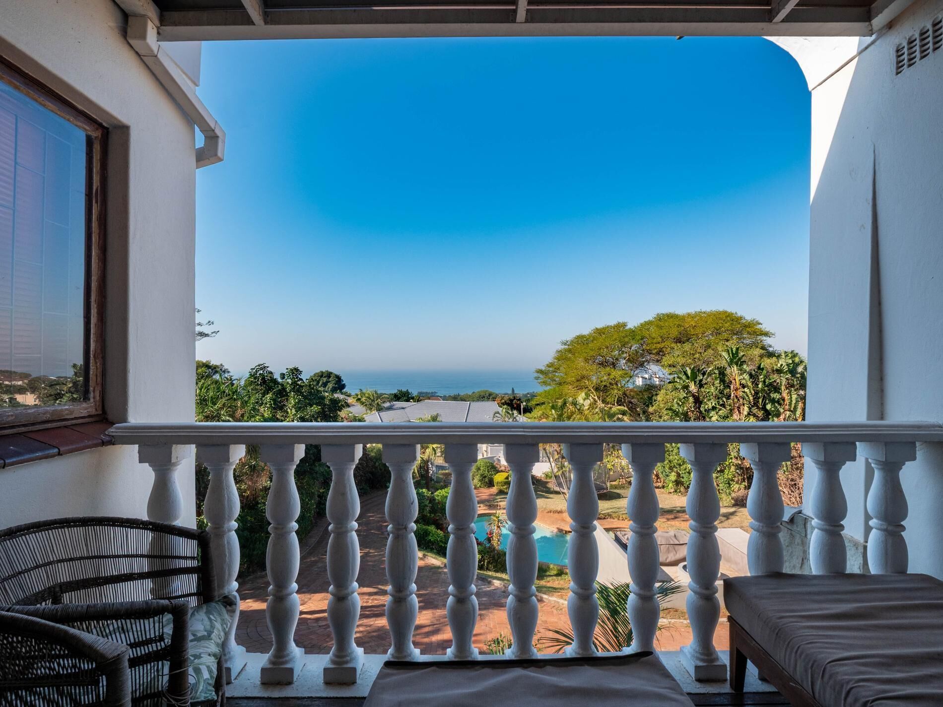 Un balcon doté d’une balustrade blanche, d’une chaise en osier, d’un banc rembourré et offrant une vue sur une piscine et la verdure.