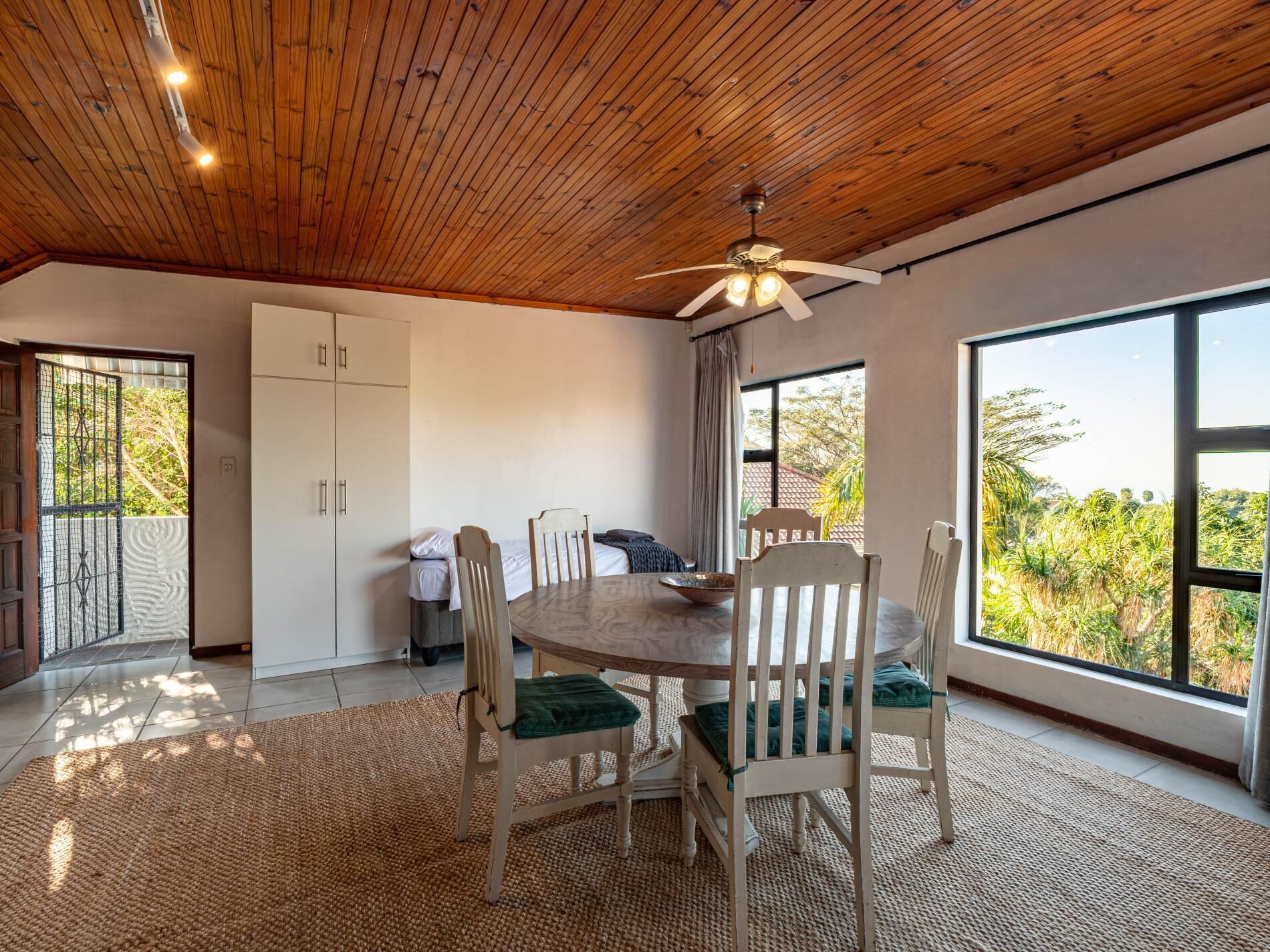 A dining area with a round table and chairs, a ceiling fan, and large windows with a view of greenery.