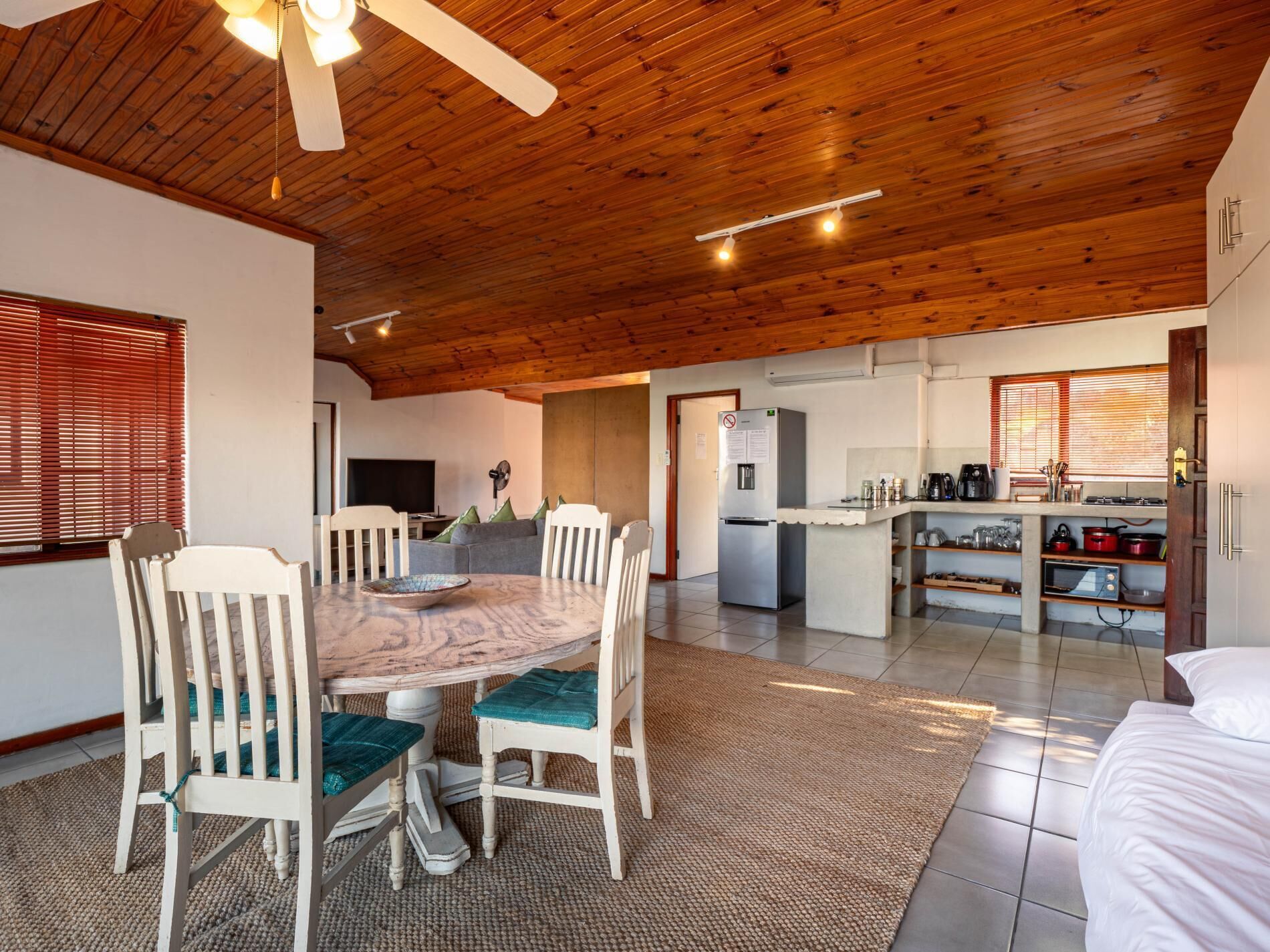 Un vaste espace de vie doté d’un plafond en bois, d’une table à manger avec des chaises, d’un coin cuisine avec un réfrigérateur et d’une grande fenêtre avec des stores.