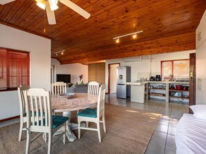 Un espacio habitable amplio con techo de madera, una mesa de comedor con sillas, una zona de cocina con refrigerador y un ventanal grande con persianas.