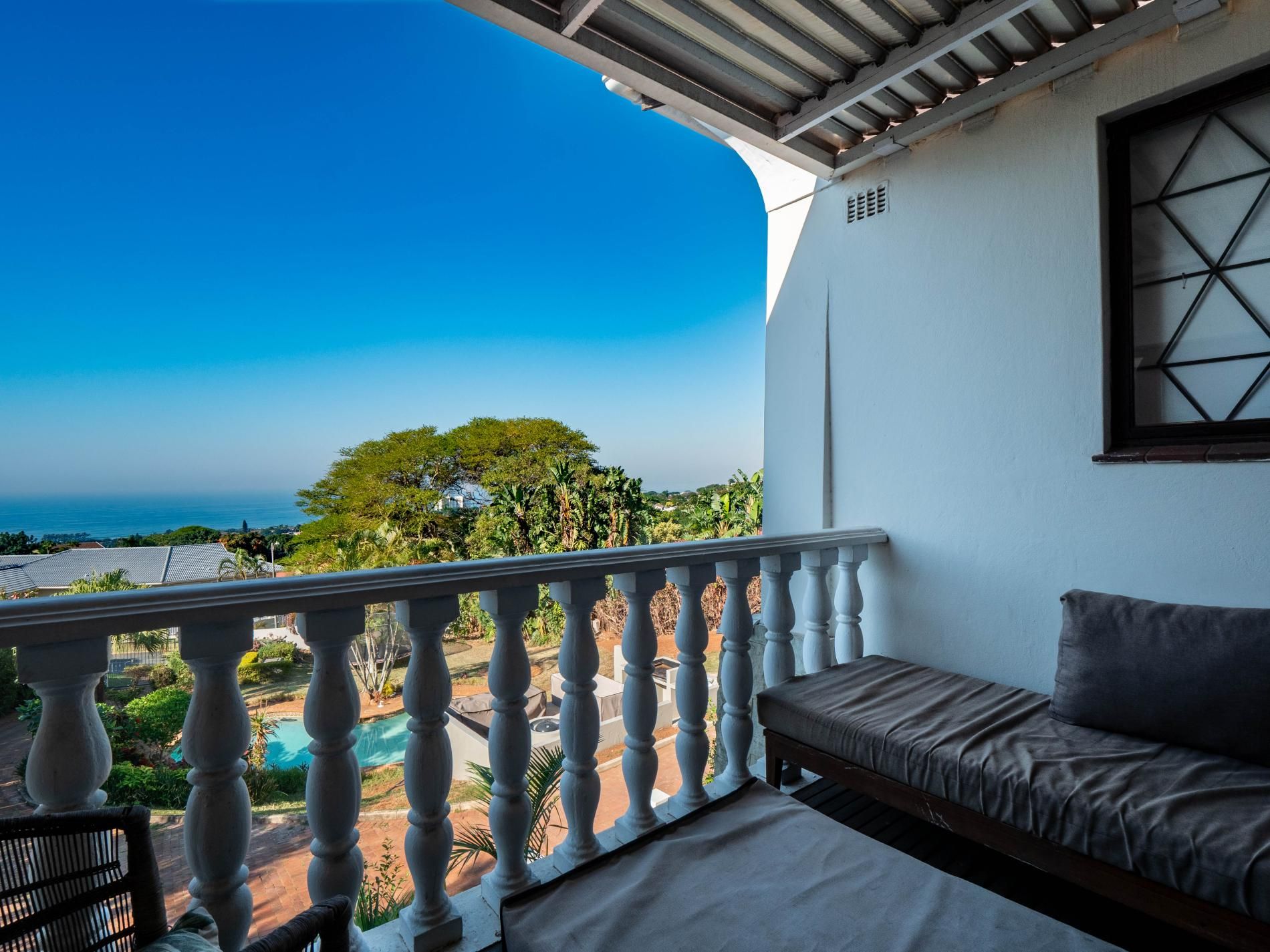 A balcony with a view of the ocean and a tree, a white wall, and a window with a geometric pattern.