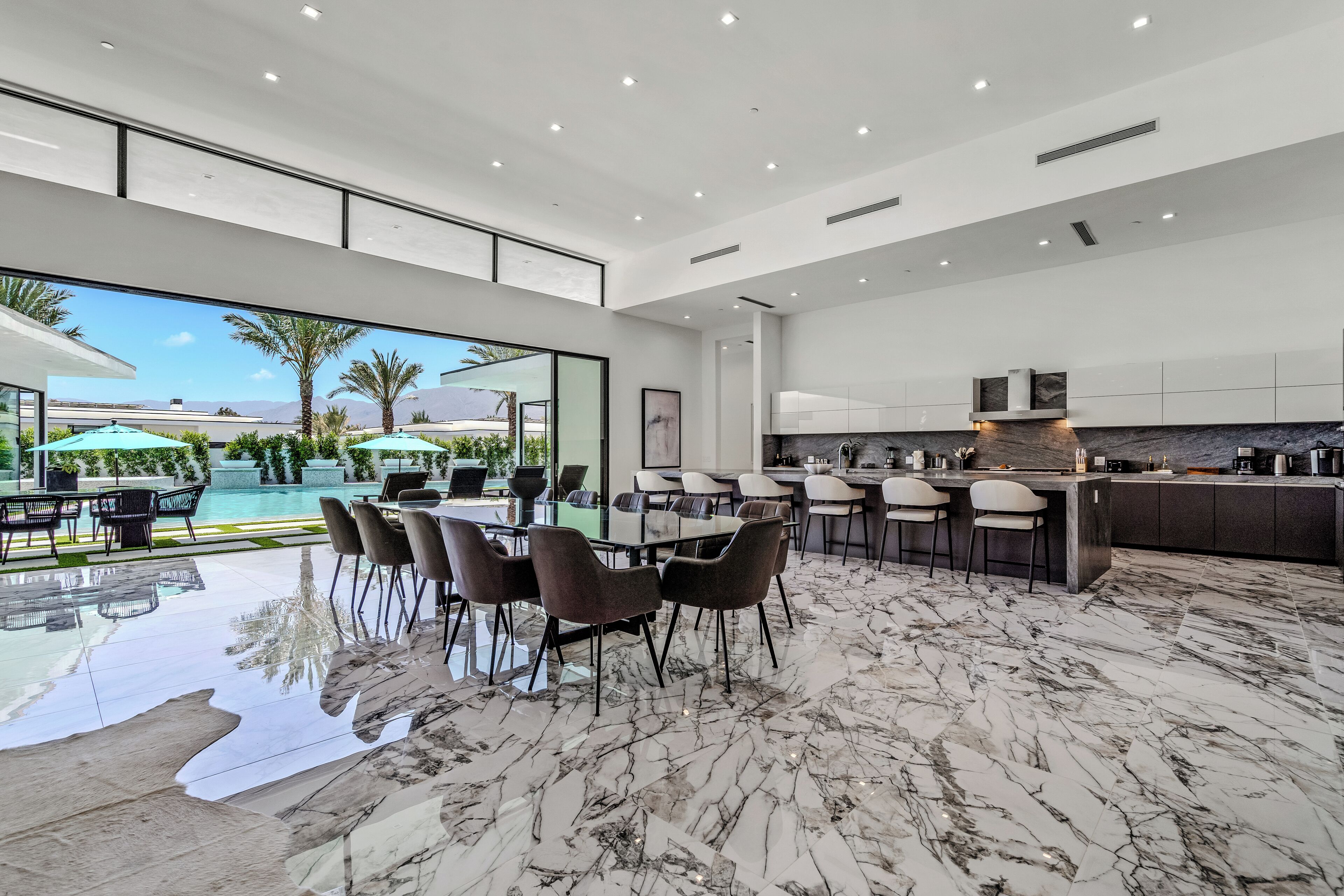 A spacious, modern kitchen with a marble floor, a dining area with a glass table and chairs, and a view of a pool and palm trees.