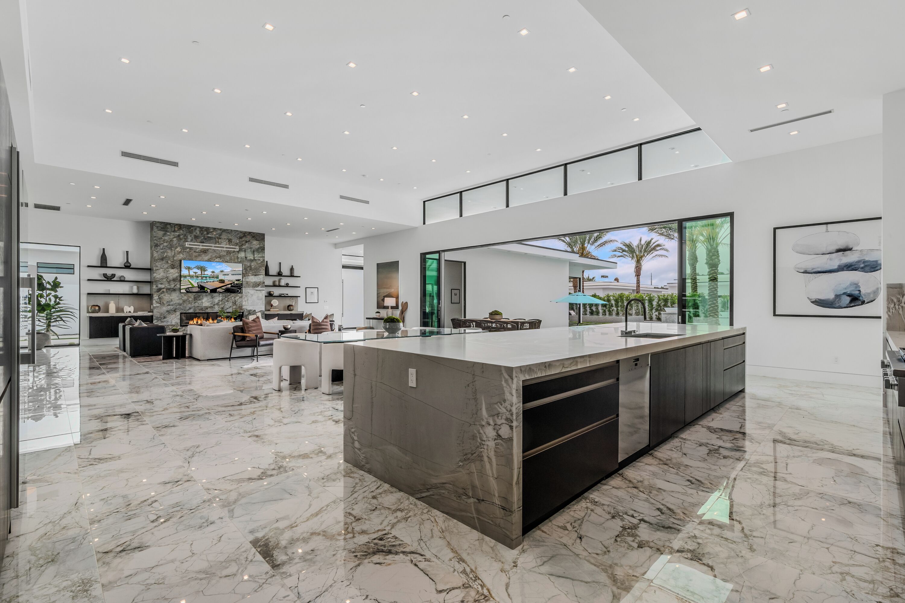 A spacious, modern kitchen with a large island, sleek cabinetry, and a large window offering a view of palm trees.