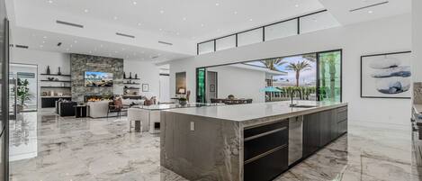 A spacious, modern kitchen with a large island, sleek cabinetry, and a large window offering a view of palm trees.