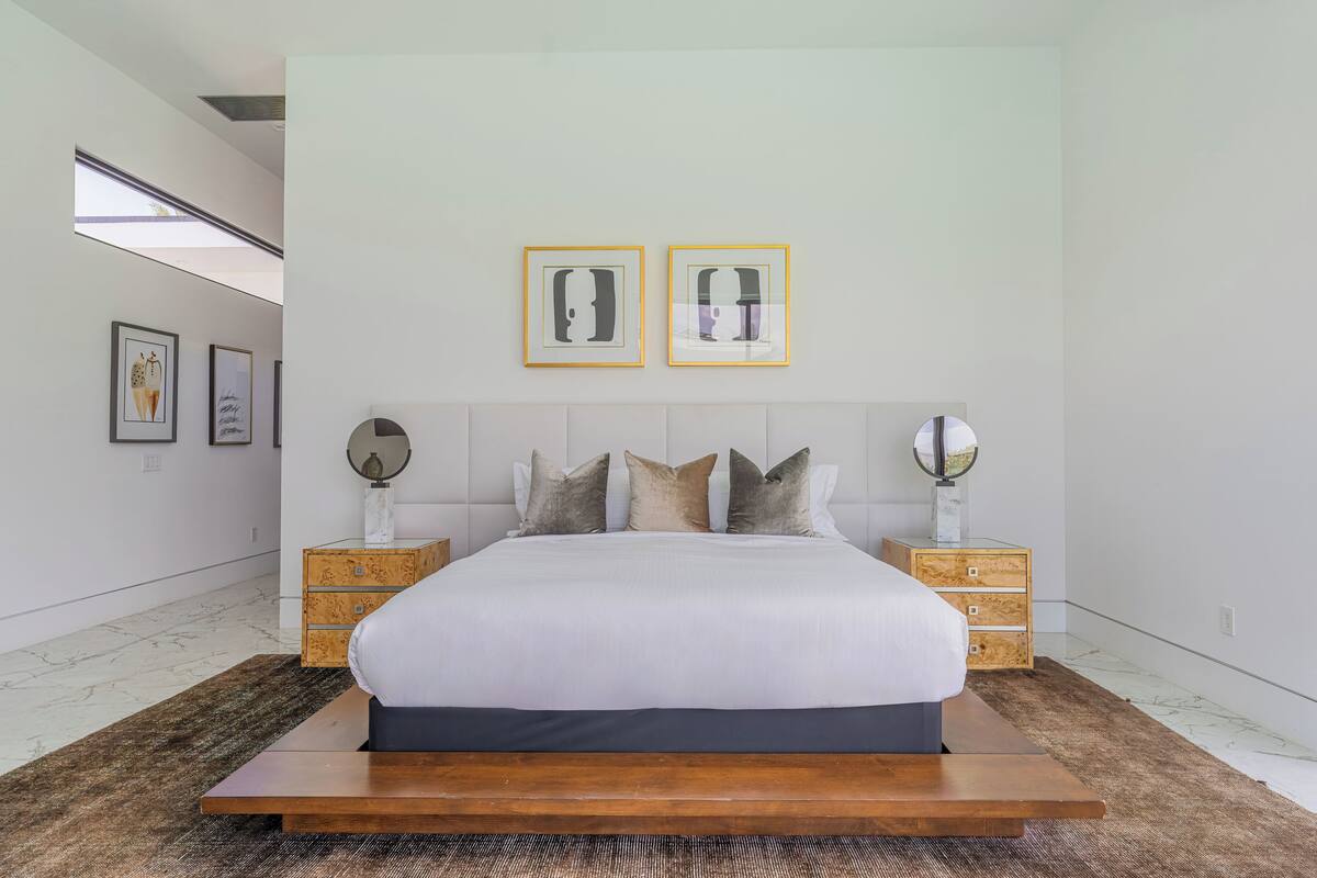 Een moderne slaapkamer met een groot bed, een houten hoofdeinde, twee nachtkastjes met lampen en een kunstwerk in een lijst aan de muur.