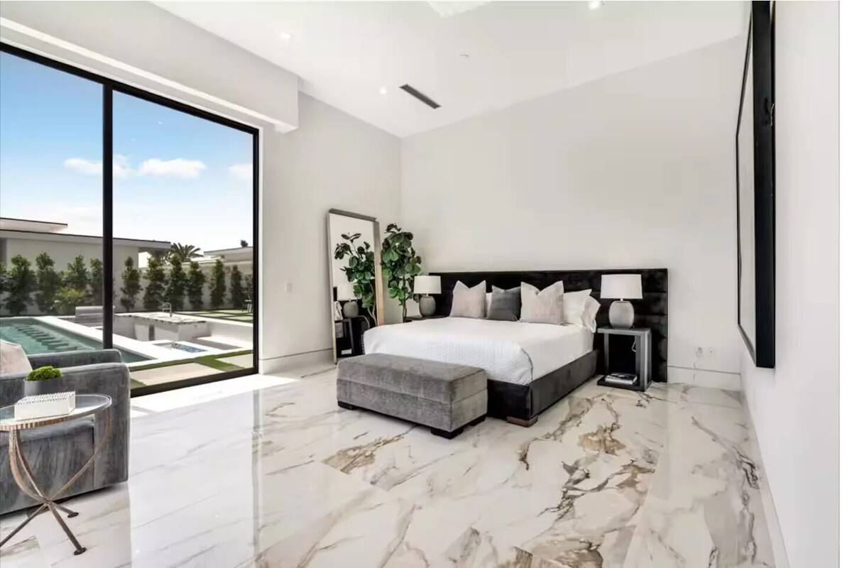 Een moderne slaapkamer met een groot bed, een zithoek met een fauteuil en tafel, en uitzicht op een zwembad en groen.