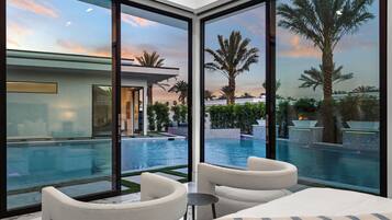 A modern hotel room with a large glass window overlooking a swimming pool and palm trees, featuring a sofa and a small table.