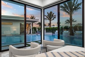 Habitación de hotel moderna con un amplio ventanal que da a una piscina y palmeras, que incluye un sofá y una mesita.