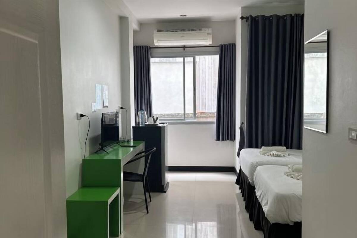 A hotel room with a desk, two beds, a window with curtains, and an air conditioning unit.