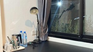 A room with a desk, a lamp, a window with curtains, and a view of trees outside. - Big Elephant Hotel (Yasothon)