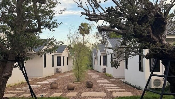 A pathway leading to a row of white cottages with dark shutters, surrounded by trees and a stone walkway. - Big Elephant Hotel (Yasothon)