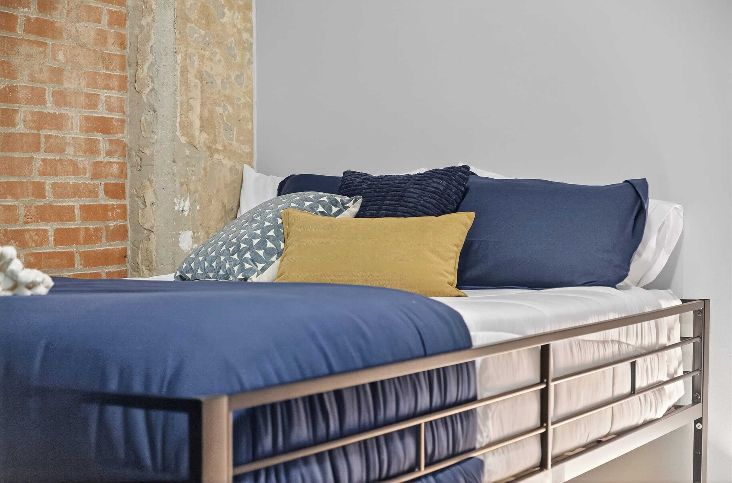 Una cama con estructura metálica, ropa de cama azul y varias almohadas en una habitación con pared de ladrillo y techo blanco.