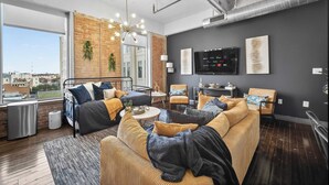 A modern living room with a sofa, armchairs, a coffee table, and a TV. There is a large window providing a view of the city. - The Bentley Collection - Urban Luxe Loft, Free Valet, Pool & 24/7 Gym (Dallas)
