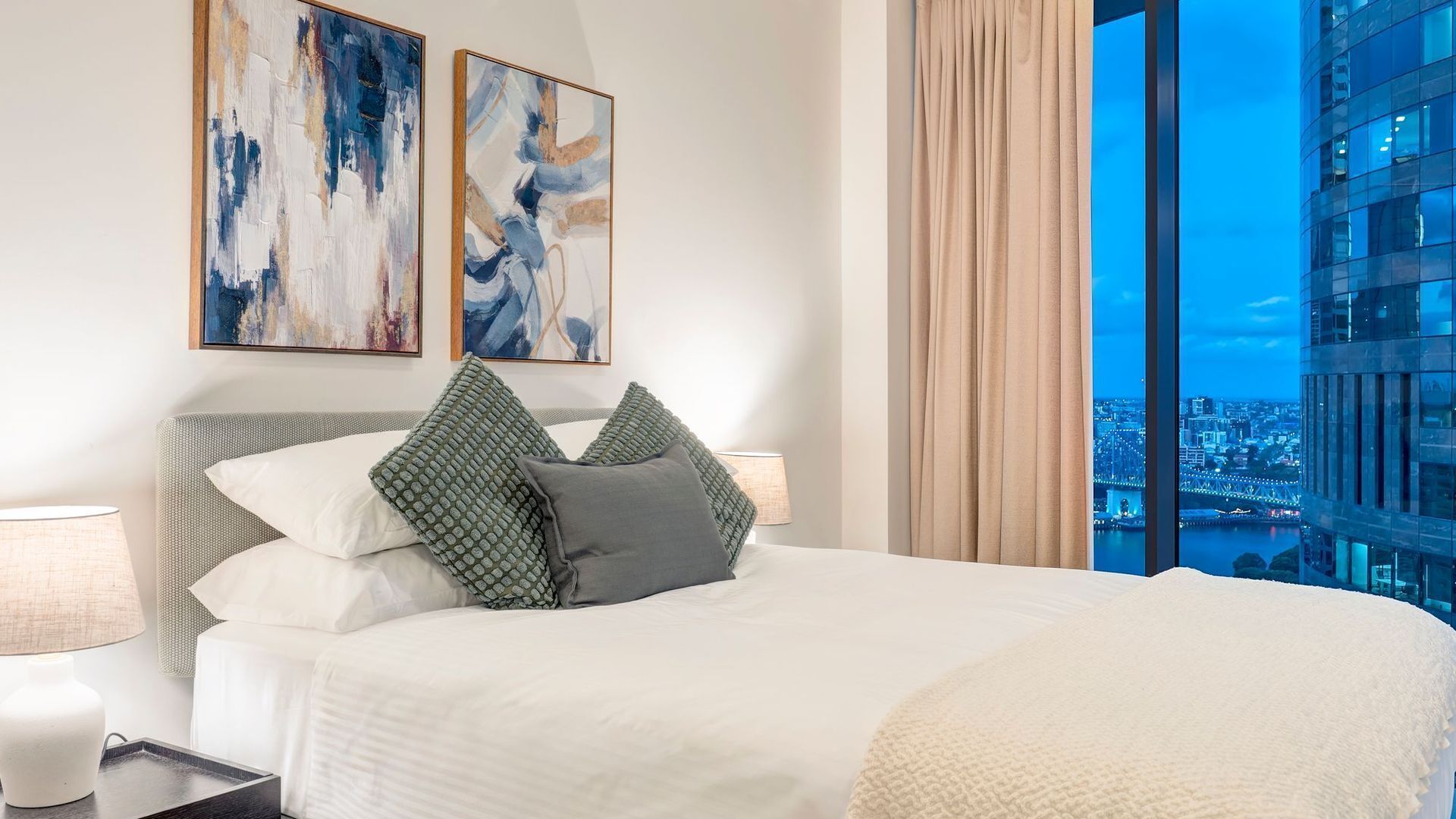 Quarto de hotel com uma cama grande, duas pinturas abstratas, vista para a cidade e um edifício moderno ao fundo.