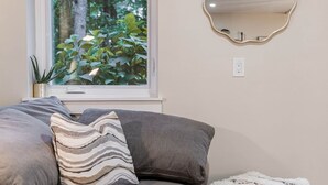 A living room with a gray sofa, a striped pillow, a fluffy ottoman, a round mirror, and a window with a view of greenery. - The Luna Lodge - close to old Orchard beach (Saco)