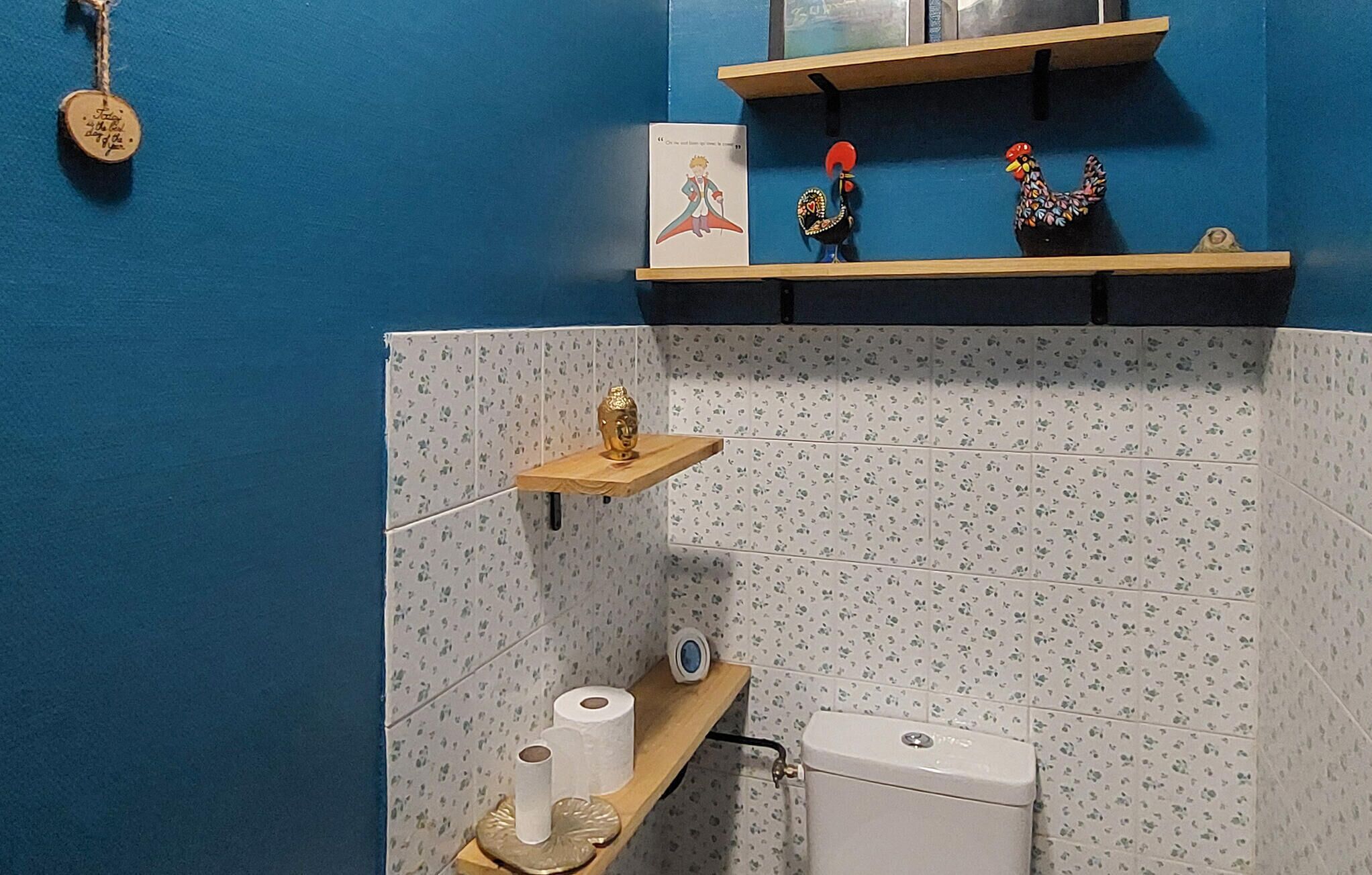 A bathroom with blue walls, a toilet, a shelf with a Buddha statue, and a small shelf with a rooster and a book.