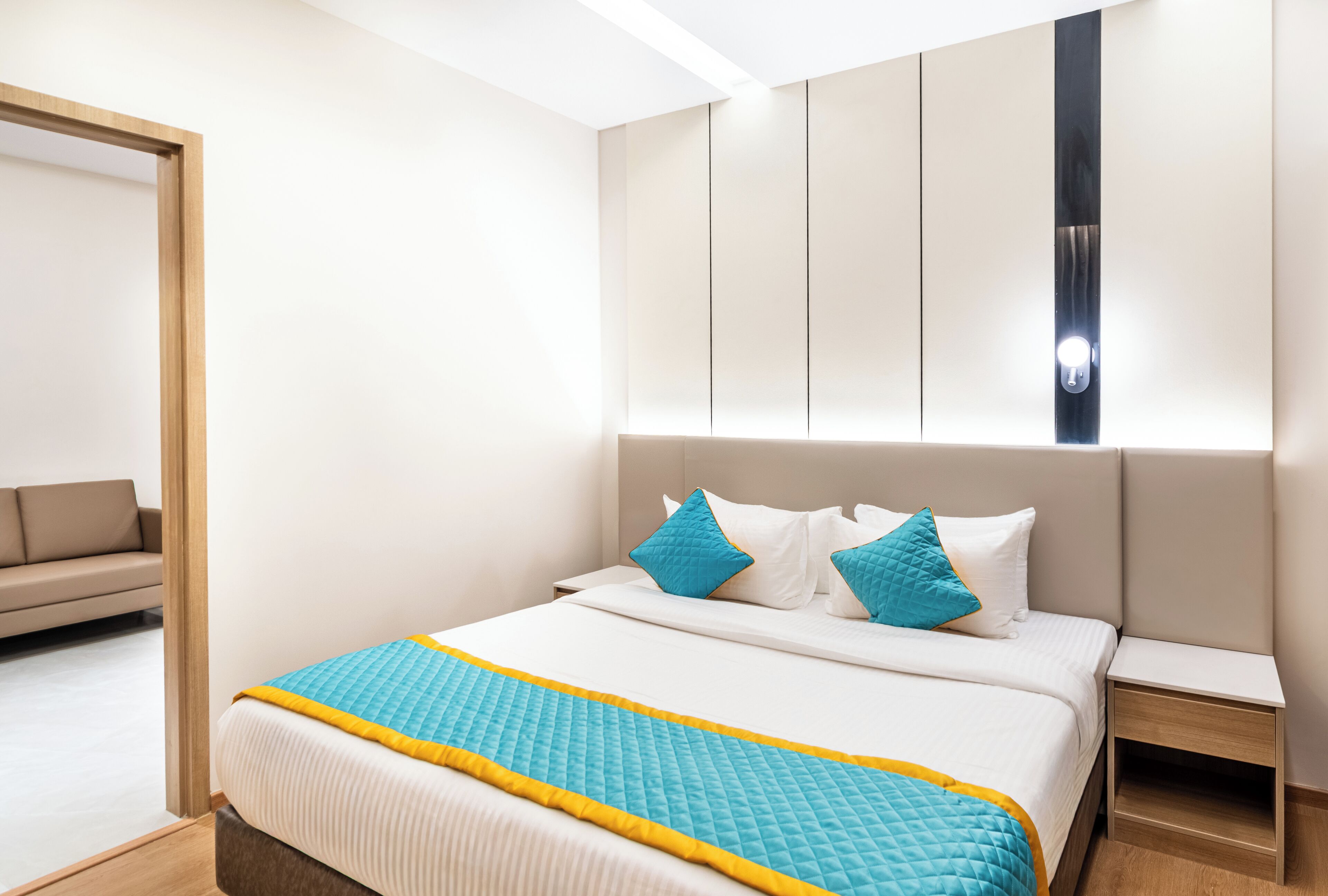 A modern hotel room with a large bed, a nightstand, a sofa, and a wall-mounted light fixture.