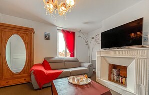 A living room with a grey sofa, a wooden cabinet with a mirror, a red throw blanket, a tray with cups and a flower, a fireplace with books, and a TV on the wall.