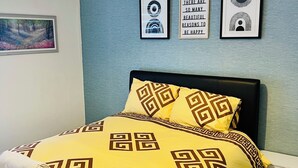 A neatly made bed with a yellow and brown patterned bedspread, a white towel, and a black headboard. There are three framed pictures on the wall, one of which has a quote.