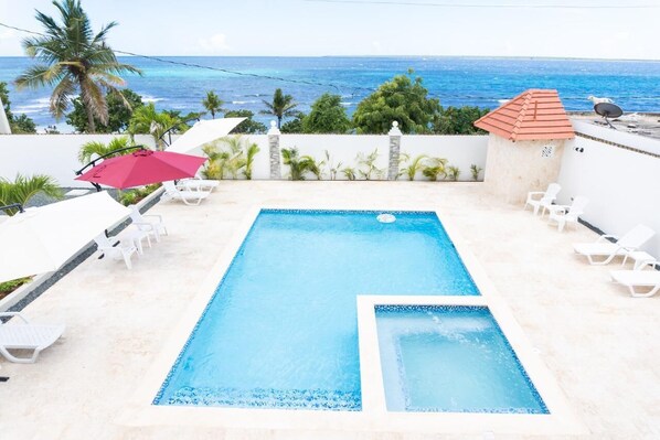 A rectangular swimming pool with a small adjacent hot tub, surrounded by white lounge chairs and a red umbrella.