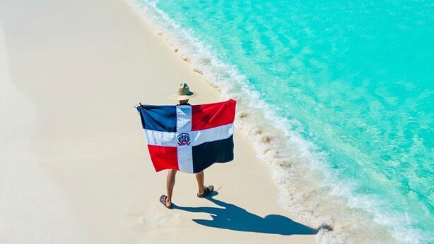 Une personne tenant un drapeau sur une plage, avec une eau turquoise et cristalline.