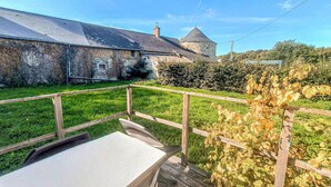 Eine Holzterrasse mit einem Tisch, ein Steinhaus mit einem Turm und ein Garten mit Weinreben.