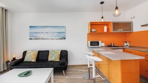 A compact living space with a kitchenette, a sofa with colorful cushions, a painting of a seascape, and a white table with a remote control. - Amadores Top Views apartment (Puerto Rico de Gran Canaria)