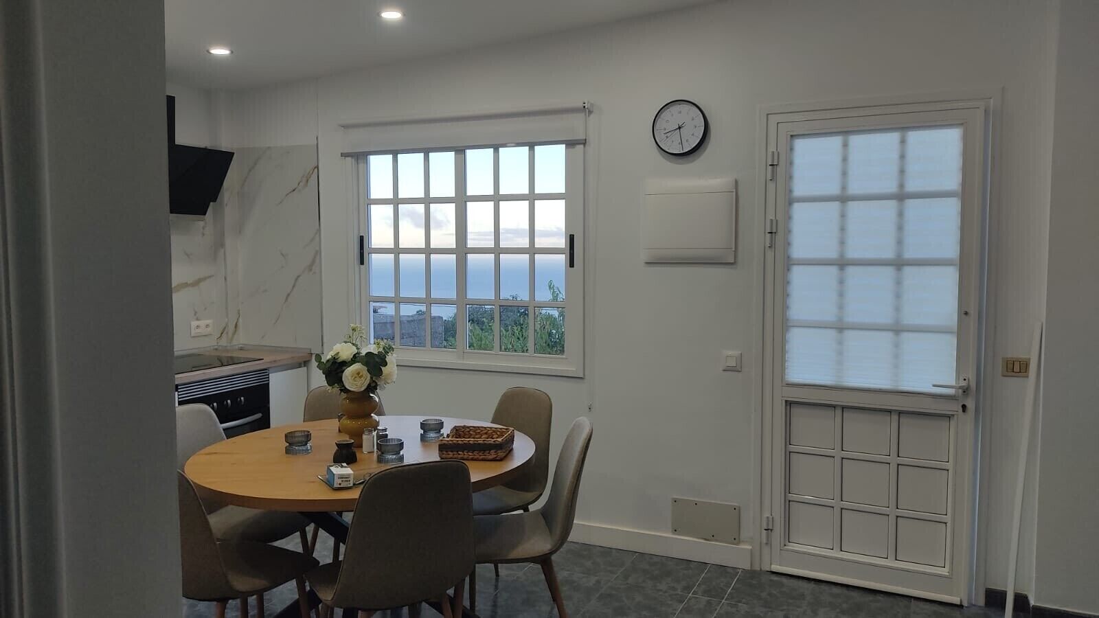 A dining area with a round table, chairs, a vase of flowers, and a view of the sea through a large window.