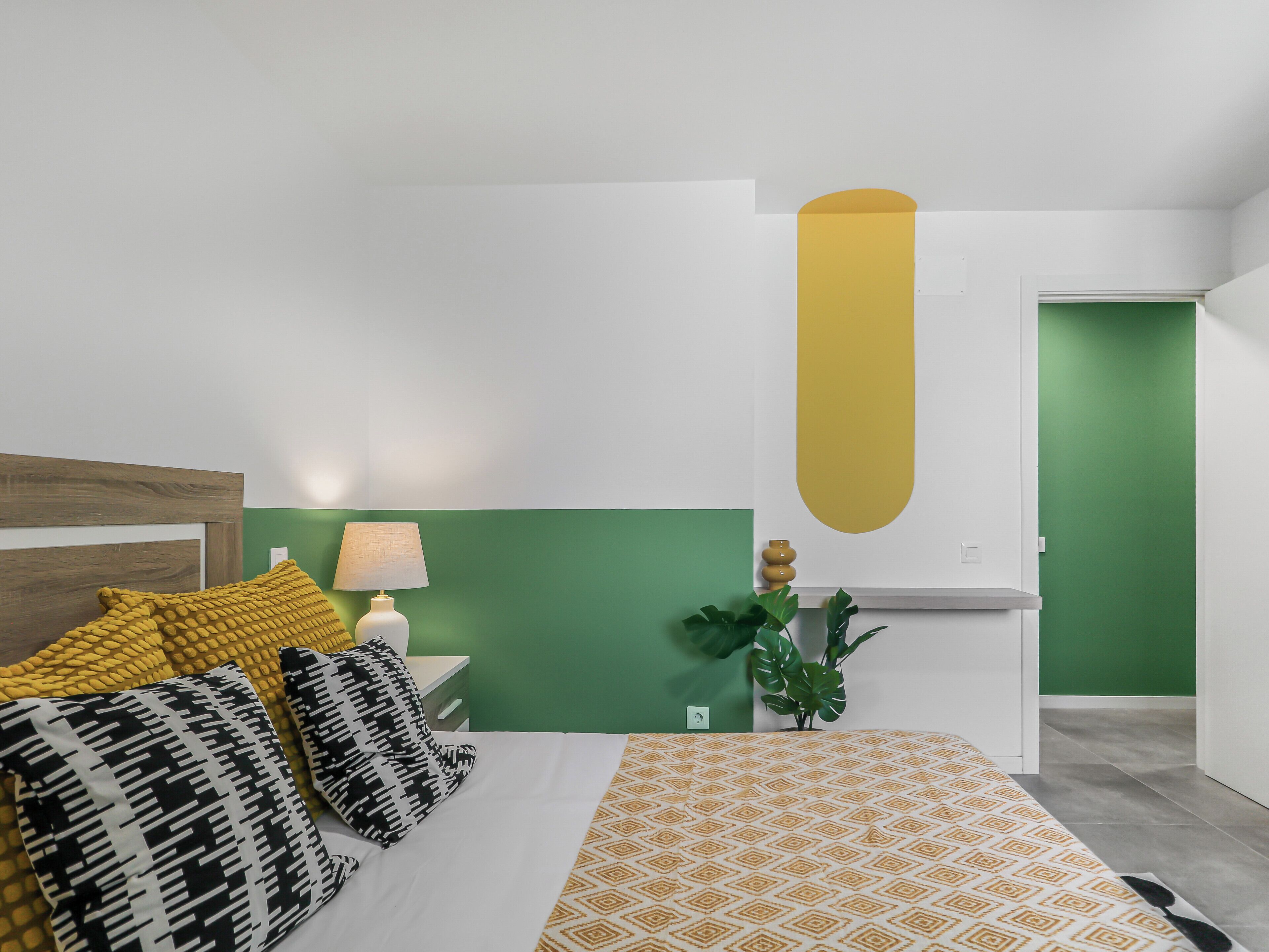 Un dormitorio con cama, almohadas, lámpara de noche, pared verde y una puerta.