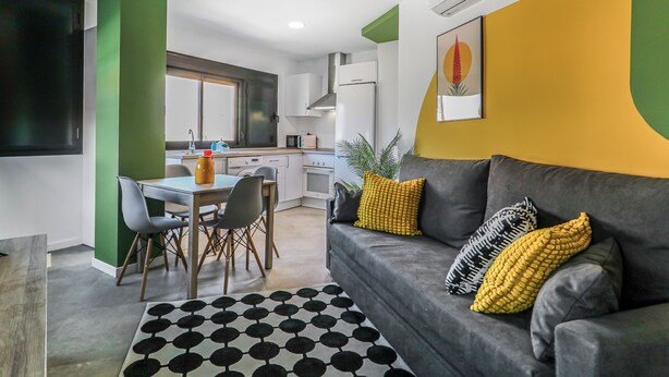 Een moderne keuken met witte kasten, een eethoek met een tafel en stoelen, en een woonkamer met een bank en kussens.