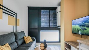 Ein modernes Wohnzimmer mit grauem Sofa, einem Couchtisch, einem an der Wand montierten Fernseher und einem Fenster mit Jalousien.