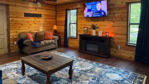 A cozy cabin-style room with a wooden interior, a television mounted on the wall, a sofa with cushions, a coffee table, and a fireplace. - Ishki Creek Cabin ~Your peaceful cabin getaway (Broken Bow)