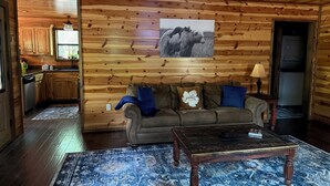 A cozy cabin interior with a wooden interior, a sofa with decorative pillows, a coffee table, and a kitchen area visible through an open door. - Ishki Creek Cabin ~Your peaceful cabin getaway (Broken Bow)