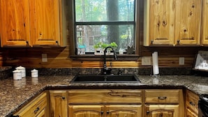 A kitchen with wooden cabinets, granite countertops, a window with a view of trees, and a rug on the floor. - Ishki Creek Cabin ~Your peaceful cabin getaway (Broken Bow)