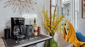 A coffee station with a coffee machine, a milk frother, and a coffee mug. There is a vase with yellow flowers and a decorative mirror on the wall.