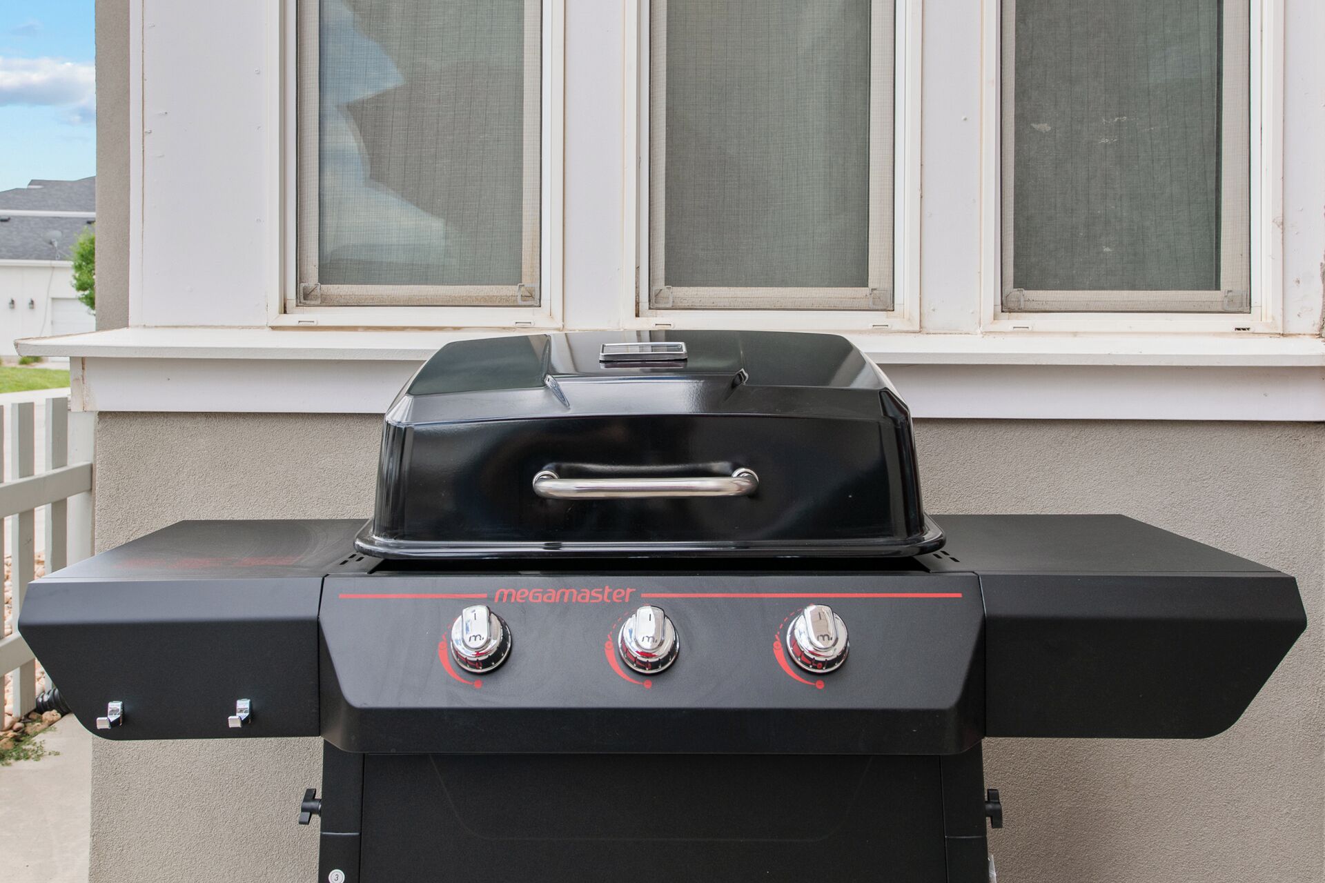 A black outdoor grill with three control knobs and a handle, placed on a balcony with a white railing and a window in the background.