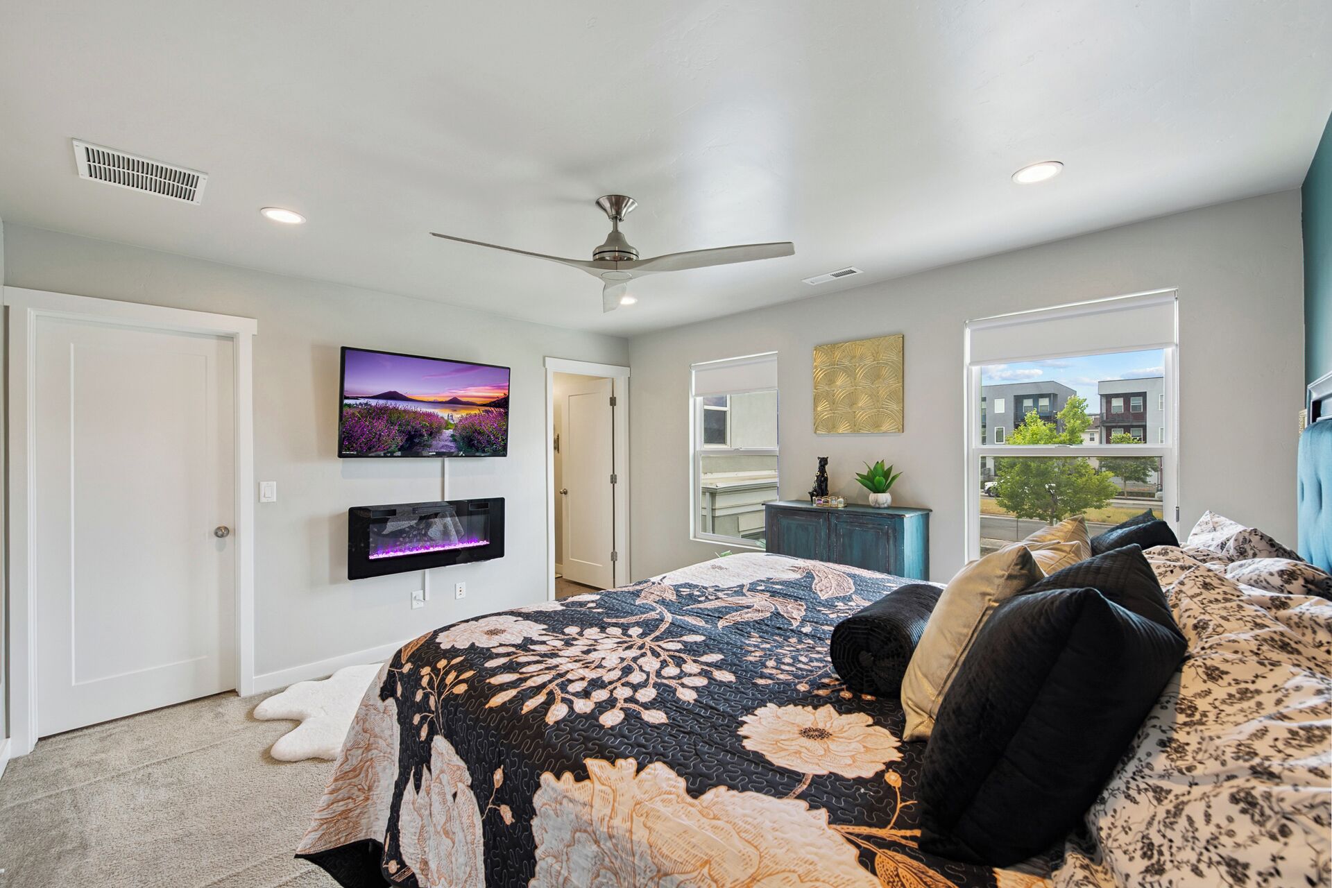 Una camera da letto con un letto, una televisione, una ventola a soffitto, una finestra e vista sugli edifici esterni.