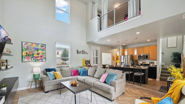 A spacious living room with a grey sofa, a coffee table, and a colorful painting. A kitchen is visible in the background with wooden cabinets and modern appliances.