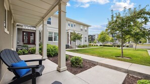 A covered patio with a bench and a tree, a well-maintained lawn, and a row of townhouses in the background.
