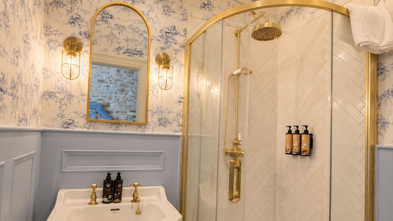 A bathroom with a white sink, gold fixtures, a shower with a gold showerhead, and a mirror with a gold frame.