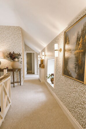 A hallway with patterned wallpaper, a painting of a forest, and a staircase leading to another room.