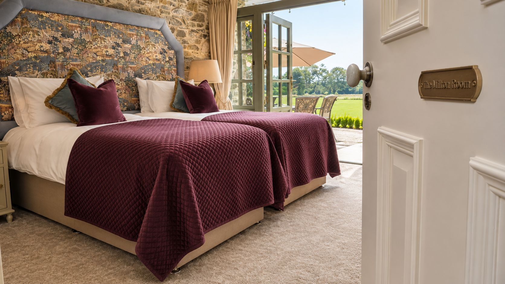 A double bed with a quilted cover, a stone headboard, and a view of the outdoors.