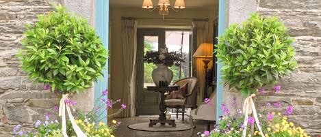 A stone building entrance with a blue door, two potted plants, a chandelier, and a table with chairs inside.