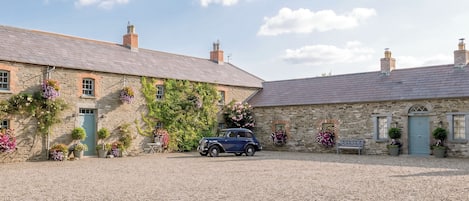 A stone building with a car parked in front, surrounded by gravel and flowering plants.
