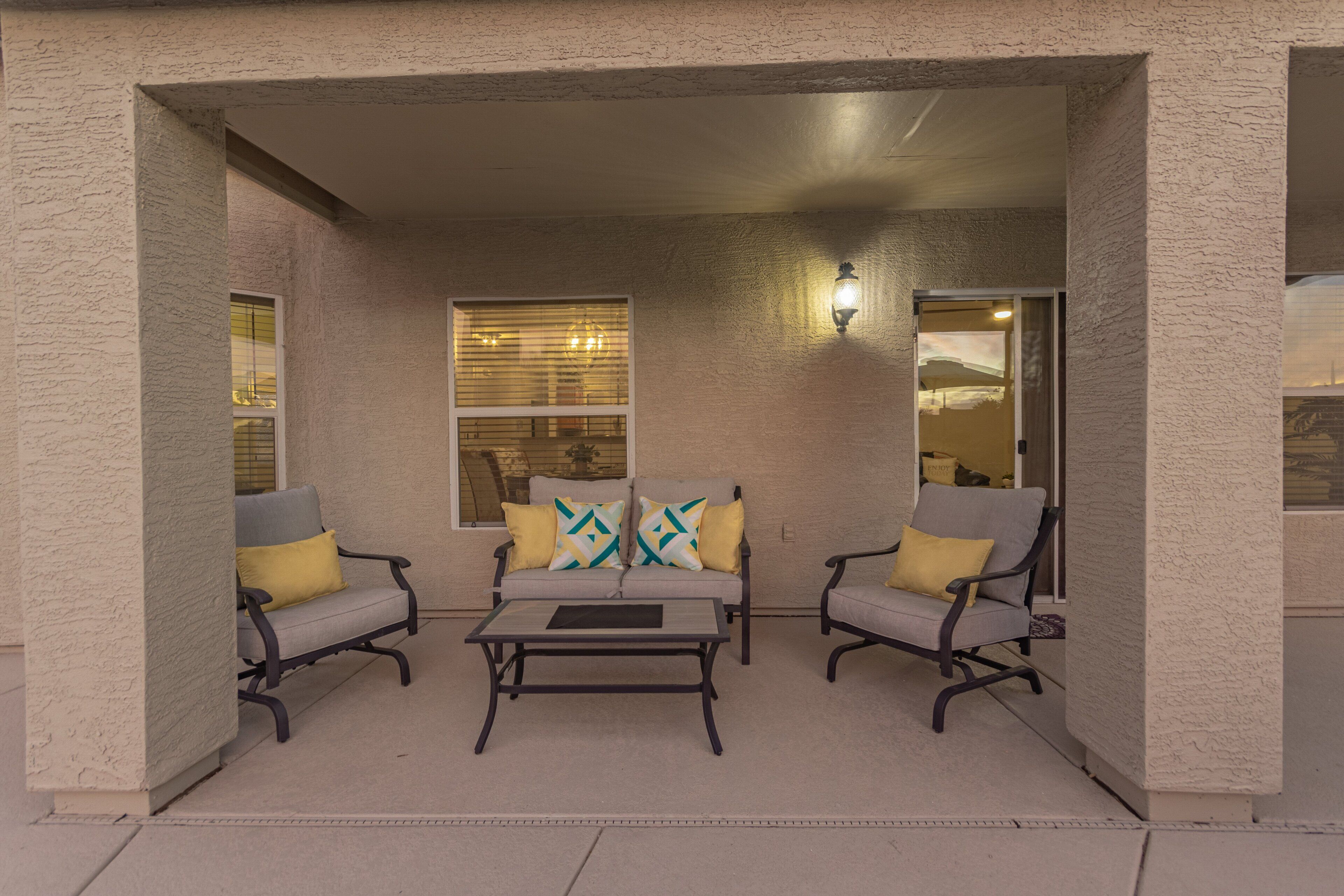 An outdoor patio with two grey armchairs, a small table, and a sofa with patterned cushions.