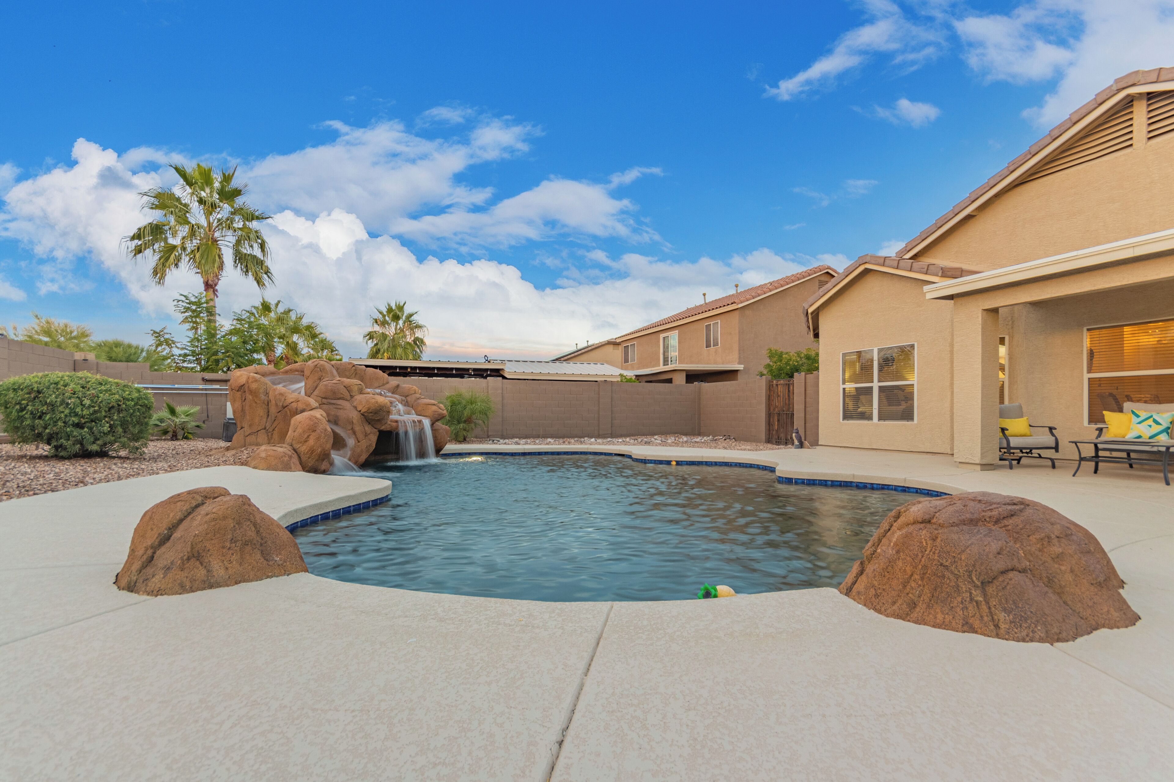 A swimming pool with a waterfall feature, surrounded by large rocks and a patio area with outdoor furniture.