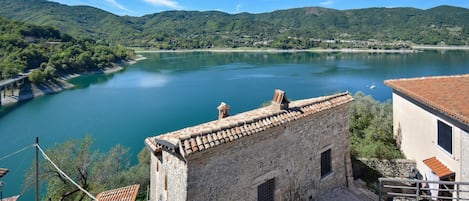 A stone building with a tiled roof overlooking a calm lake, surrounded by lush green hills.