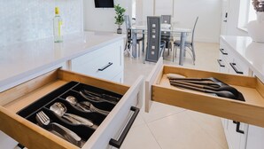 A modern kitchen with white cabinets, a wooden drawer with utensils, and a dining area with a table and chairs. - FLORIDA PARADISE / 5 MIN TO DOWNTOWN / 3BDR/6BEDS (Fort Myers)